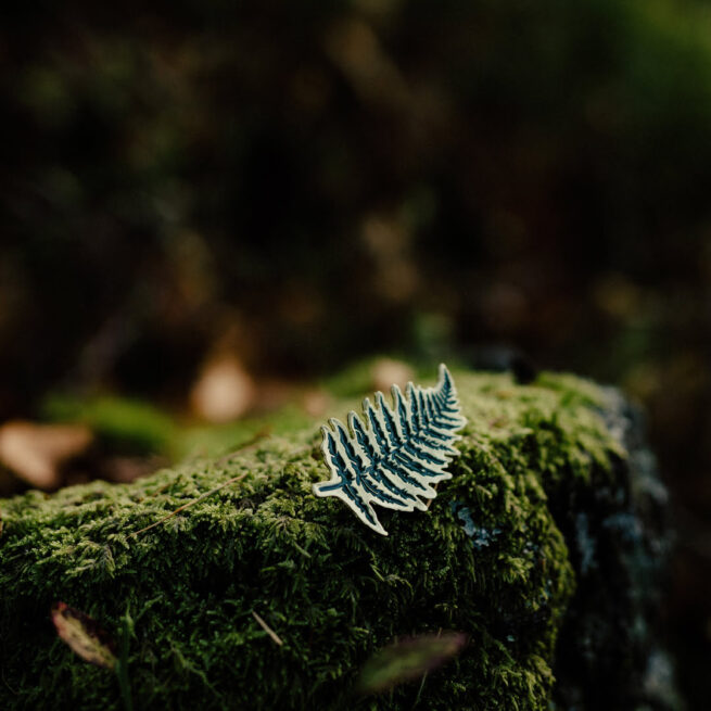 Metalowe ręcznie emaliowane pinsy w kształcie liścia paproci leżące kamieniu obrośniętym mchem w otoczeniu lasu od producenta pinów emaliowanych od Pinswear