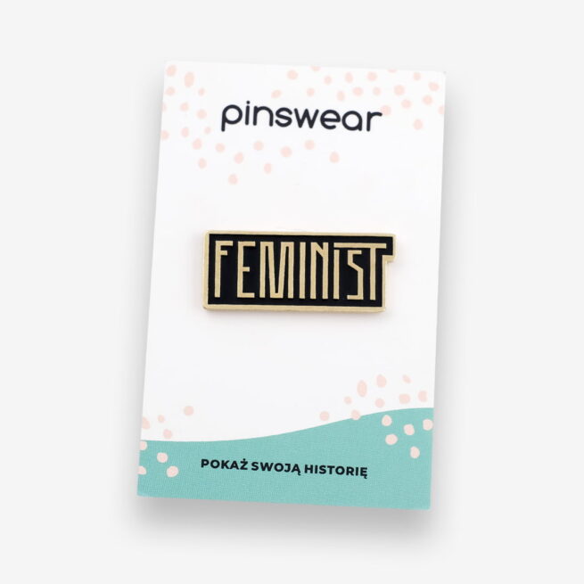 Przypinki do ubrań napis złoty napis feminist na czarnym tle. Pins w złotym wykończeniu wpięty na personalizowany kartonik z logo pinswear.